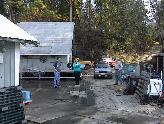 Three people gathered in a yard near a workshop building with cars and trees